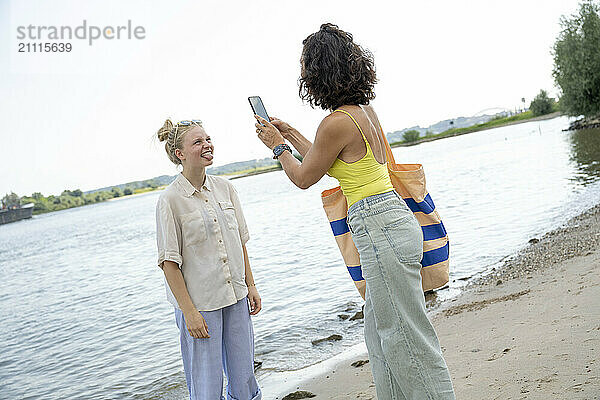 A woman takes a photo of her friend by a lake with a smartphone on a sunny day.