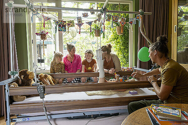 Family members gather around a hospital bed decorated with toys and balloons  spending time with a patient in a home-like environment.