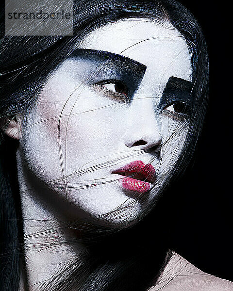 Dramatic portrait of a woman with bold black and white facial makeup and vivid red lips  gazing off to the side on a dark background.