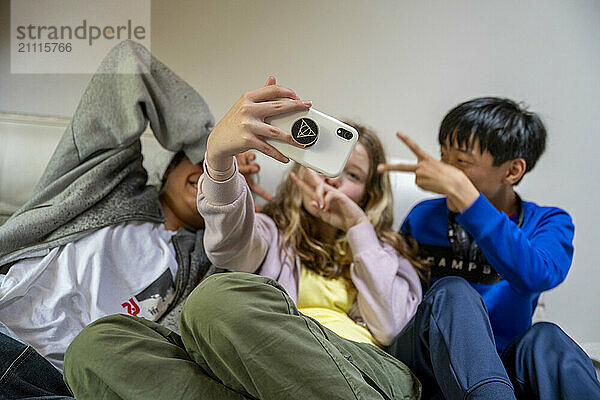 Three kids take a selfie together  one covering their face with a hoodie  another making a peace sign  and the third smiling at the camera.