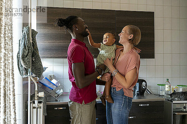 A joyful family moment as parents share a smile while lifting their baby in the kitchen.