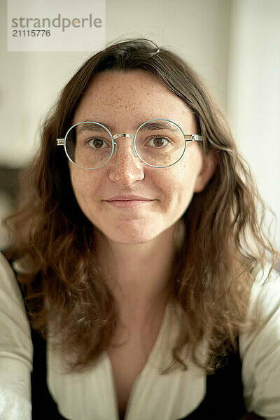 Smiling young adult woman with round glasses and wavy hair in a formal white blouse against a soft-focus background.