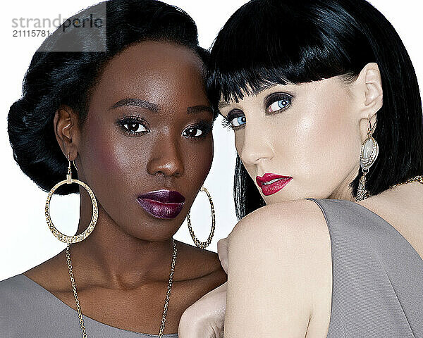 Two women with striking makeup and large hoop earrings pose confidently against a white background.