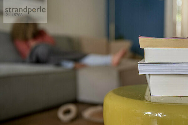 Stacked books on a yellow table with a blurred woman reading in the background on a gray sofa