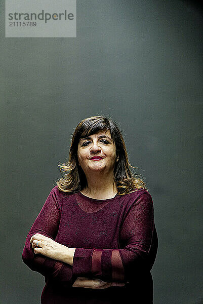 Confident woman in a burgundy top standing with arms crossed against a gray background.