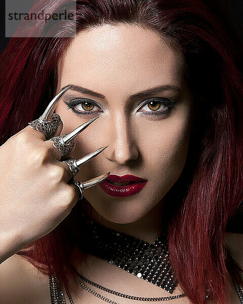 Red-haired woman with striking makeup wearing claw rings and looking at the camera against a black background.