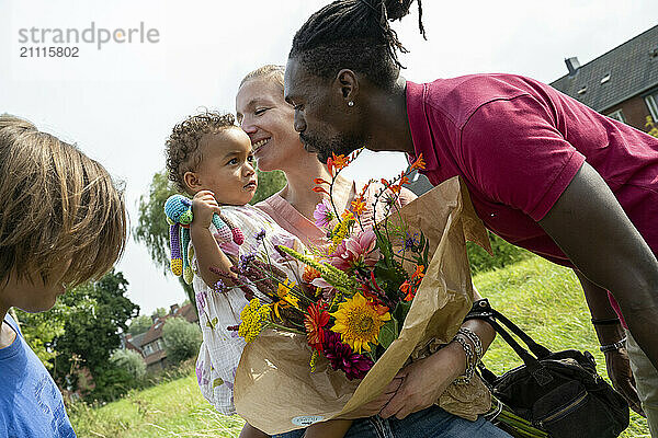 A smiling man in a red shirt kisses a toddler held by a woman with a bouquet of flowers as another child watches.