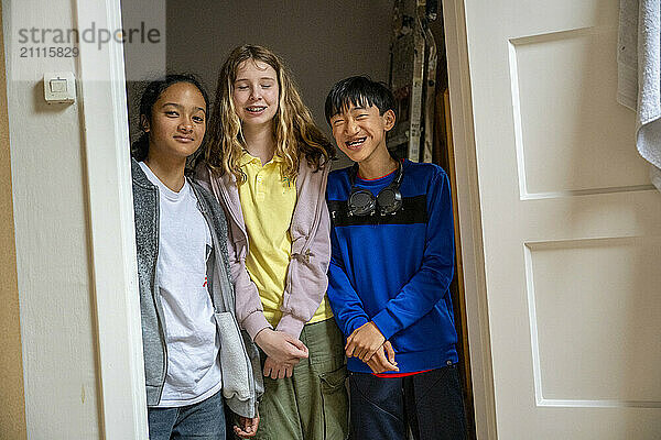 Three cheerful children standing in a doorway  smiling and posing for a photo with natural daylight illuminating their faces.