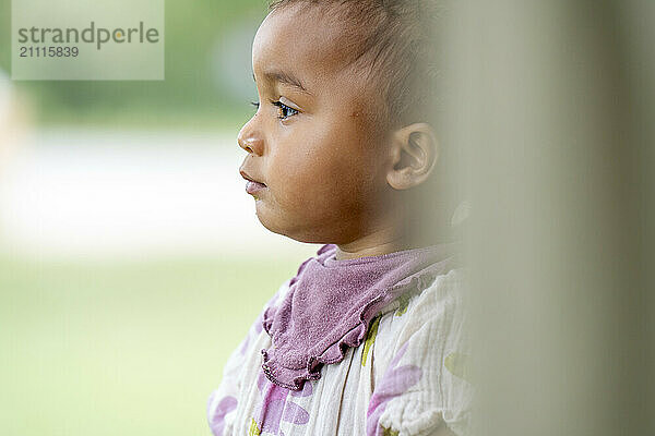 Side profile of a toddler with a thoughtful expression  dressed in a bib  against a blurred natural background.