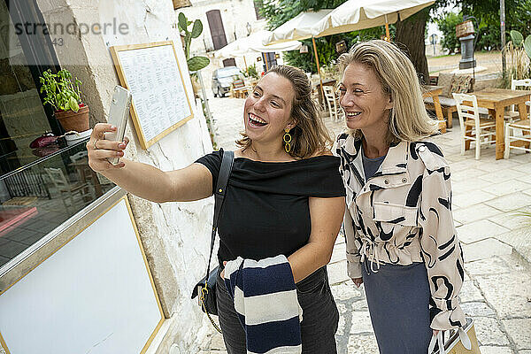 Two women taking a selfie together on a city street with a restaurant terrace in the background.