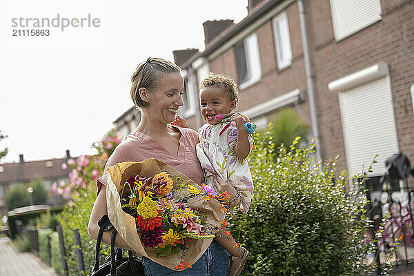 A smiling woman carrying her cheerful toddler and a bouquet of flowers while walking outdoors.