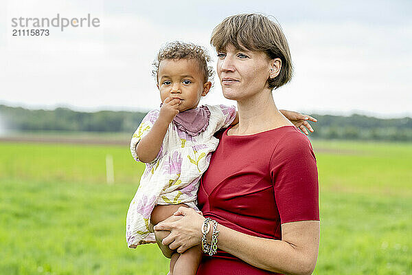 Woman in red top holding a young child outdoors with a field in the background.