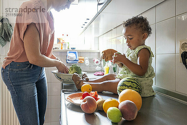 A young child sits on a kitchen counter with fruits nearby as an adult female prepares food.