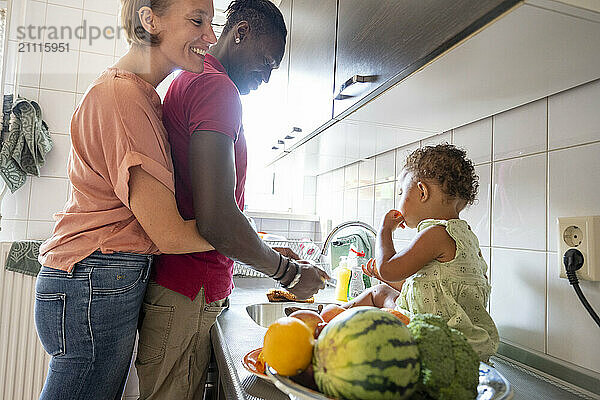 A happy family in the kitchen with a toddler sitting on the counter surrounded by fruit as the parents lovingly watch over.