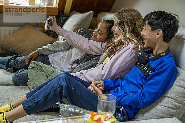 Three friends lounging on a couch take a selfie together with a smartphone  smiling and enjoying each other's company in a cozy living room.