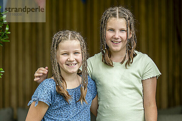 Two smiling girls with braided hair standing together in front of a wooden background.