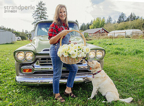 Smiling woman in a plaid shirt holding a basket of flowers stands next to a classic car with a yellow labrador retriever sitting beside her in a grassy field.