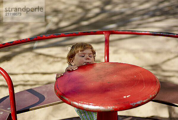 Young child closing eyes and enjoying a moment while resting head on a bright red playground spinner.