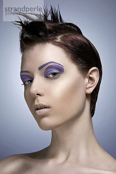 Woman with dramatic purple eyeshadow and styled hair posing against a grey background.