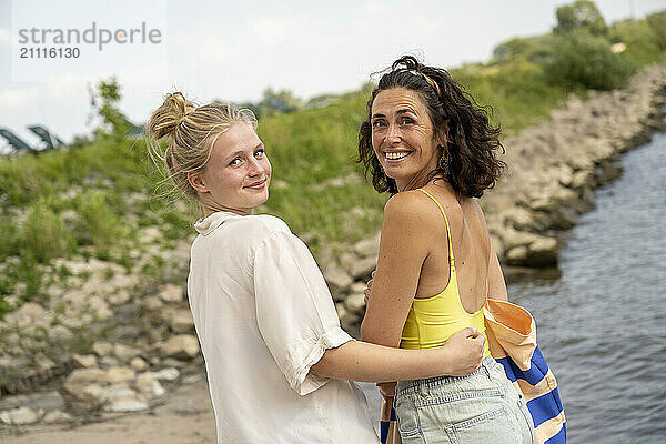 Two women smiling by a riverbank  one with her arm around the other.