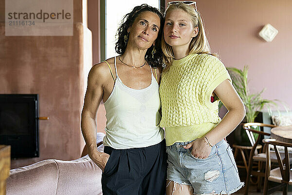 Two women standing closely together in a room with pink walls  smiling at the camera with a cozy home-like setting in the background.