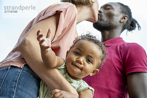 Multiracial family enjoying a moment together  with a toddler smiling at the camera while being held by parents who are sharing a kiss in the background.