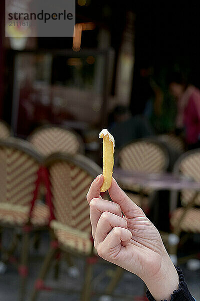 Close-up of a hand holding a french fry with a blurred outdoor cafÈ background.