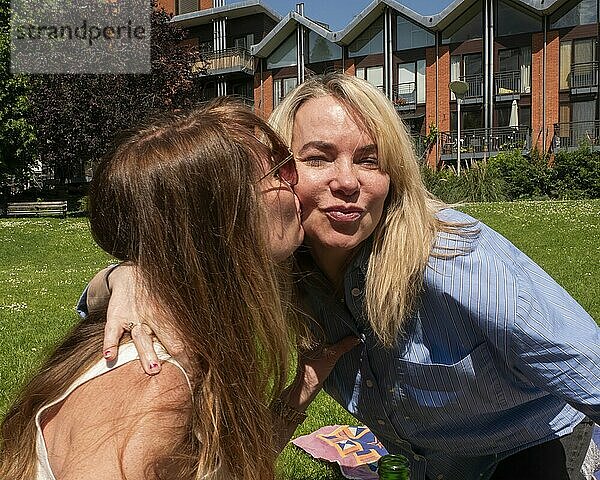 Two women sharing a friendly kiss on the cheek in a sunny park with apartment buildings in the background.