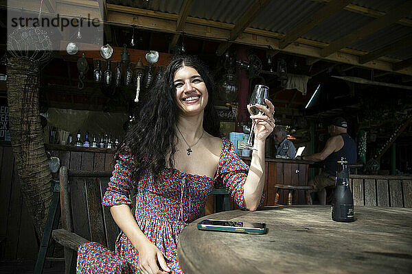 Smiling woman enjoying a drink at a rustic outdoor bar with hanging decorations and a patron in the background  Titikaveka  Rarotonga  Cook Islands