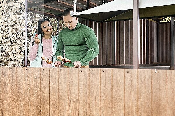 Lächelndes Mädchen in Sportkleidung und ihr gutaussehender Freund in grünem Pullover kochen Schaschlik auf einer Veranda im Freien