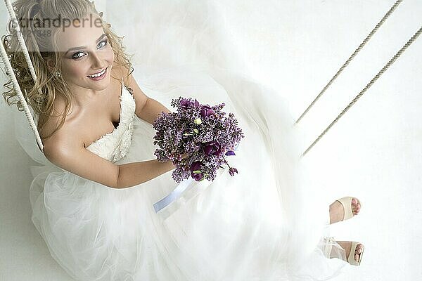 Gorgeous junge Braut posiert im Studio sitzen auf Schaukel mit einem exquisiten rosa und violetten Bouquet