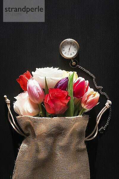 Schöner Strauß verschiedener Blumen  eingewickelt in einen rustikalen Jutesack und eine Vintage Taschenuhr  auf schwarzem Hintergrund  an einem sonnigen Tag im Frühling