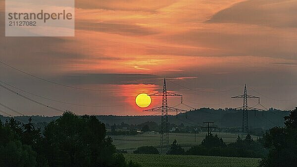 Rote Sonne  die über einem deutschen Dorf und der umgebenden grünen Natur aufgeht  und Silhouetten von Hochspannungsmasten  in der Nähe von Schwäbisch Hall  Deutschland  Europa