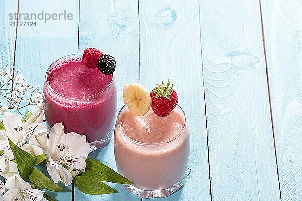 Frühstückstisch mit einem Glas frischem Bananen und Erdbeersmoothie  einem mit Himbeeren und Waldbeeren  neben weißen Blumen auf blauem Hintergrund