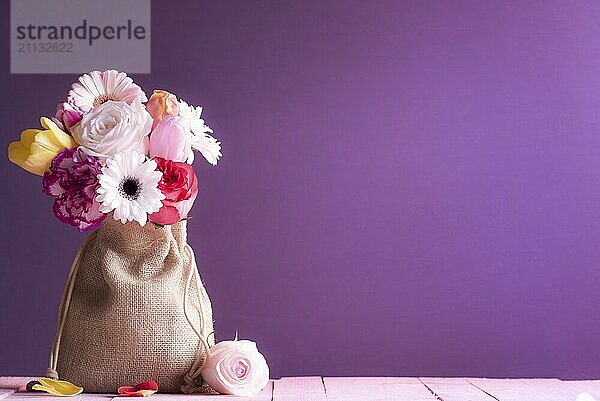 Geburtstagskarte mit einem bunten Blumenstrauß aus Rosen  Tulpen und Gerbera  eingewickelt in einen alten Leinensack  auf einem rosa Holztisch und einem lila Hintergrund