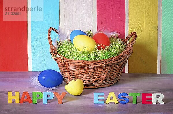 Dekorative Weidenkorb mit bemalten Eiern und die Worte frohe Ostern mit multicolor Holz Buchstaben und einem bunten Zaun im Hintergrund geschrieben