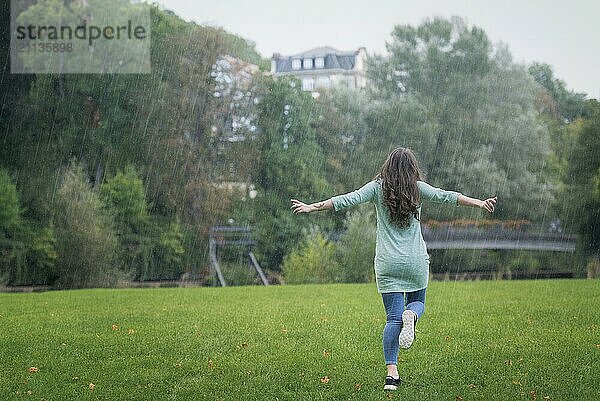 Brünette junge Frau  leger gekleidet  läuft fröhlich mit ausgebreiteten Armen durch den Regen  umgeben von grünem Gras und Bäumen  in Deutschland