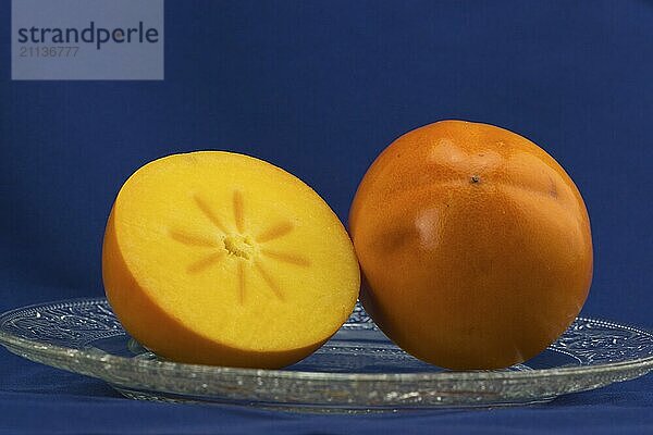 Eine halbierte Kaki  Frucht auf einem Glasteller vor blauem Hintergrund