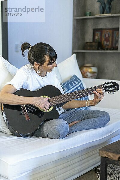 Eine erwachsene Frau  die elegant auf ihrer Gitarre klimpert  während sie sich zu Hause auf ihrem Sofa entspannt