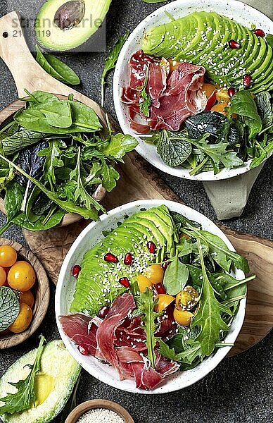 Schüssel mit wenig Kohlenhydraten. Frischer Salat mit grünem Spinat  Rucola  Avocado und Serrano Schinken in weißer Schüssel  grauer Hintergrund  Ansicht von oben