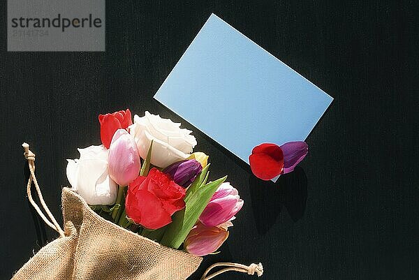 Bunter Strauß verschiedener Blumen in einem Jutesack eingewickelt und eine leere blaue Papiernotiz mit Blumenblättern darauf  auf einem schwarzen Hintergrund