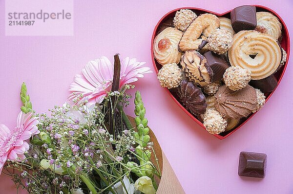 Geschenk Themenbild mit einer roten herzförmigen Schachtel voller köstlicher Süßigkeiten und einem Blumenstrauß auf einem rosa Hintergrund. Eine Idee für eine Grußkarte