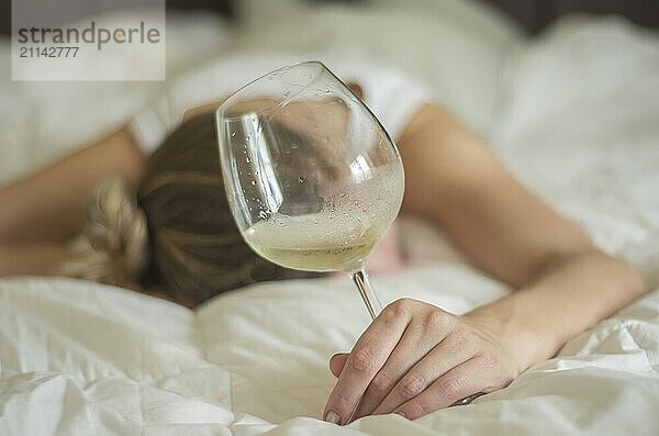 Großartiges Konzept des Alkoholmissbrauchs. Junge Frau  blondes Haar  ohnmächtig im Bett  nachdem sie zu viel Alkohol getrunken hat. Ein Glas Wein in der Hand  eine Flasche Wein