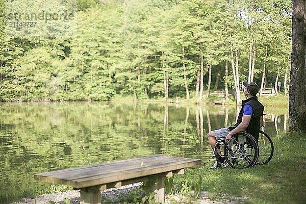 Glücklicher und junger behinderter Mann im Rollstuhl sitzend mit Blick auf einen schönen See in der Natur