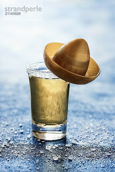 Ein Schuss Tequila mit Salz auf einem blauen Tisch mit Hintergrundbeleuchtung. Ein Glas goldener Tequila mit einem kleinen Sombrero. Traditionelles mexikanisches Getränk. Starker Alkohol