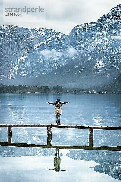 Mädchen mit ausgebreiteten Armen  das auf einem schmalen Holzdeck steht und den Blick auf die Berge der österreichischen Alpen genießt  die sich im Wasser des Hallstätter Sees spiegeln  in Österreich