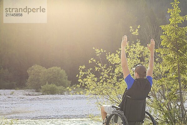 Glücklicher und fröhlicher junger Mann im Rollstuhl draußen in der Natur an einem sonnigen Sommertag mit erhobenen Händen