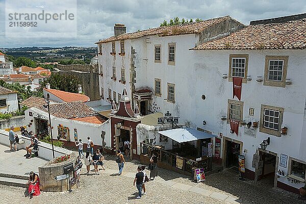 Menschen spazieren durch eine kopfsteingepflasterte historische Altstadt mit weißen Gebäuden und roten Dächern  Blick vom Miradouro Porta  Largo de Padrão  Óbidos  Obidos  Oeste  Centro  Estremadura  Portugal  Europa