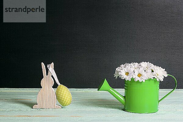 Cute white daisies arrangement in a watering can and a wooden bunny with a yellow fabric egg hanging  on a green table and a black background