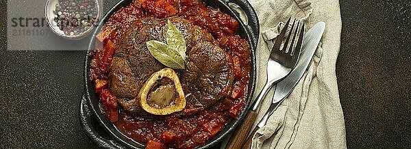 Traditionelles italienisches Gericht Ossobuco all Milanese aus geschnittener Kalbshaxe mit vegetarischer Tomatensoße  serviert in schwarzer Kasserolle von oben auf rustikalem braunem Hintergrund  Foodfotografie